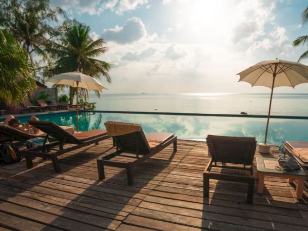 A serene poolside scene with lounge chairs, umbrellas, palm trees, and a view of a calm sea under a partly cloudy sky.