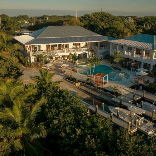 Aerial view of a tropical resort with a central pool, multiple white buildings, palm trees, and a dock by a calm canal. Top it at 140 characters, always ending the sentence.