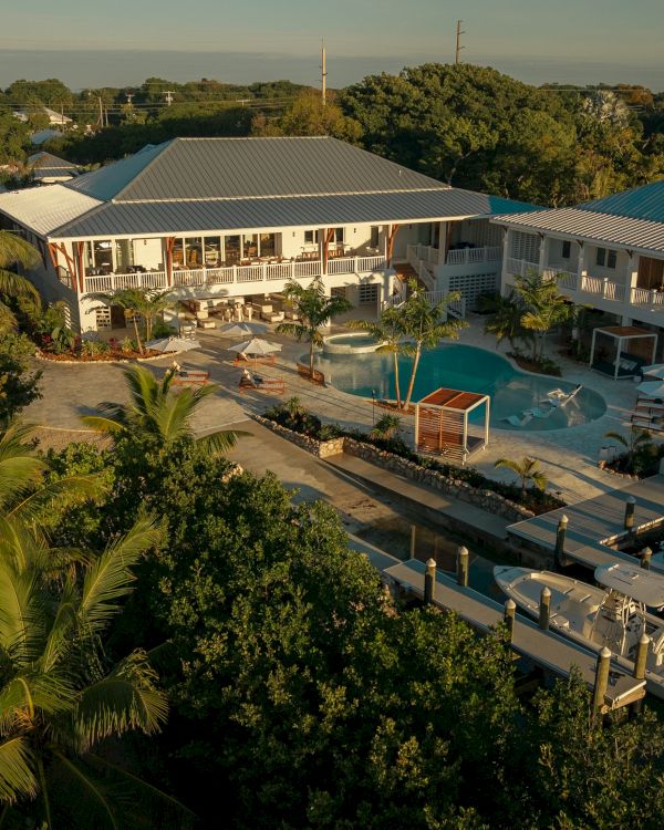 Aerial view of a tropical resort with a central pool, multiple white buildings, palm trees, and a dock by a calm canal. Top it at 140 characters, always ending the sentence.