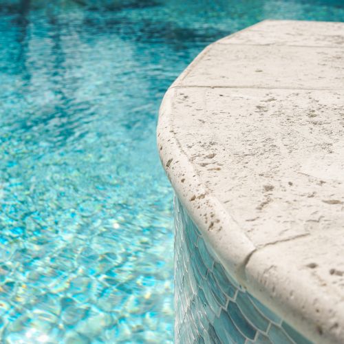 A close-up of a sparkling blue pool with water cascading over a white plaster edge, sunlight glimmering on the rippling surface.