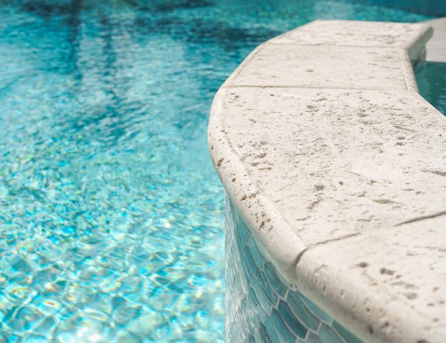 A close-up of a sparkling blue pool with water cascading over a white plaster edge, sunlight glimmering on the rippling surface.