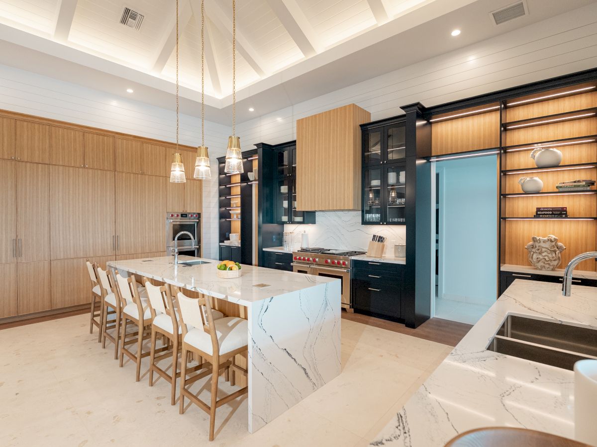 A modern kitchen with a long marble island, pendant lights, wooden bar stools, white cabinetry, black appliances, and open shelving.