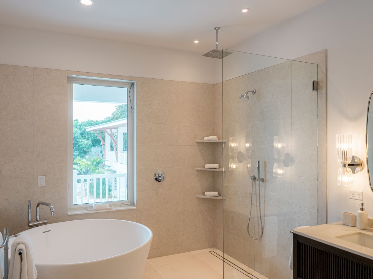 A modern bathroom with a freestanding tub, glass-walled shower, beige tiled walls, a window, and a vanity with an oval mirror inside a cozy, well-lit space.