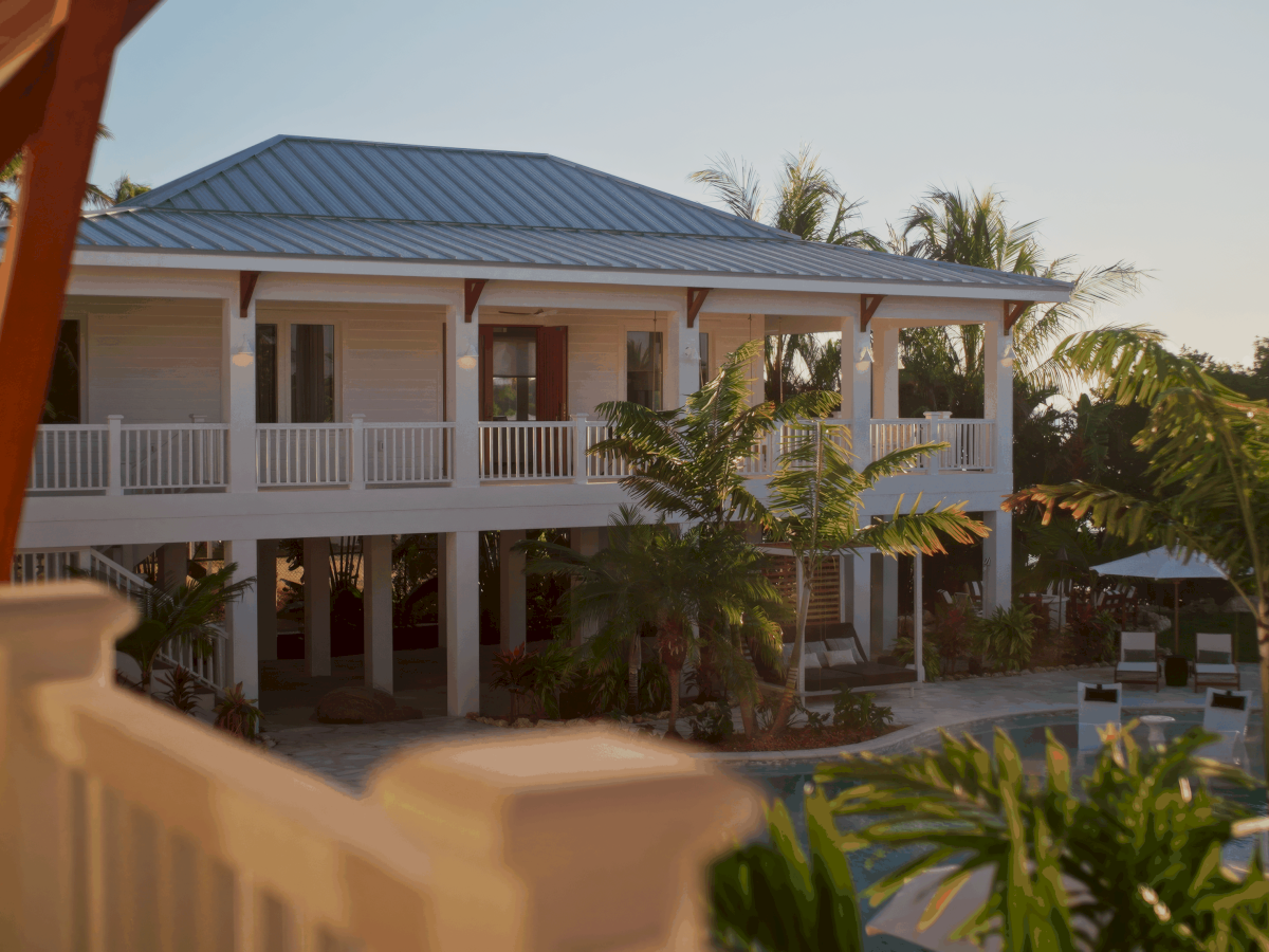 A two-story resort with white railings, tropical plants, and a pool area, set under a clear sky near a sunny veranda.