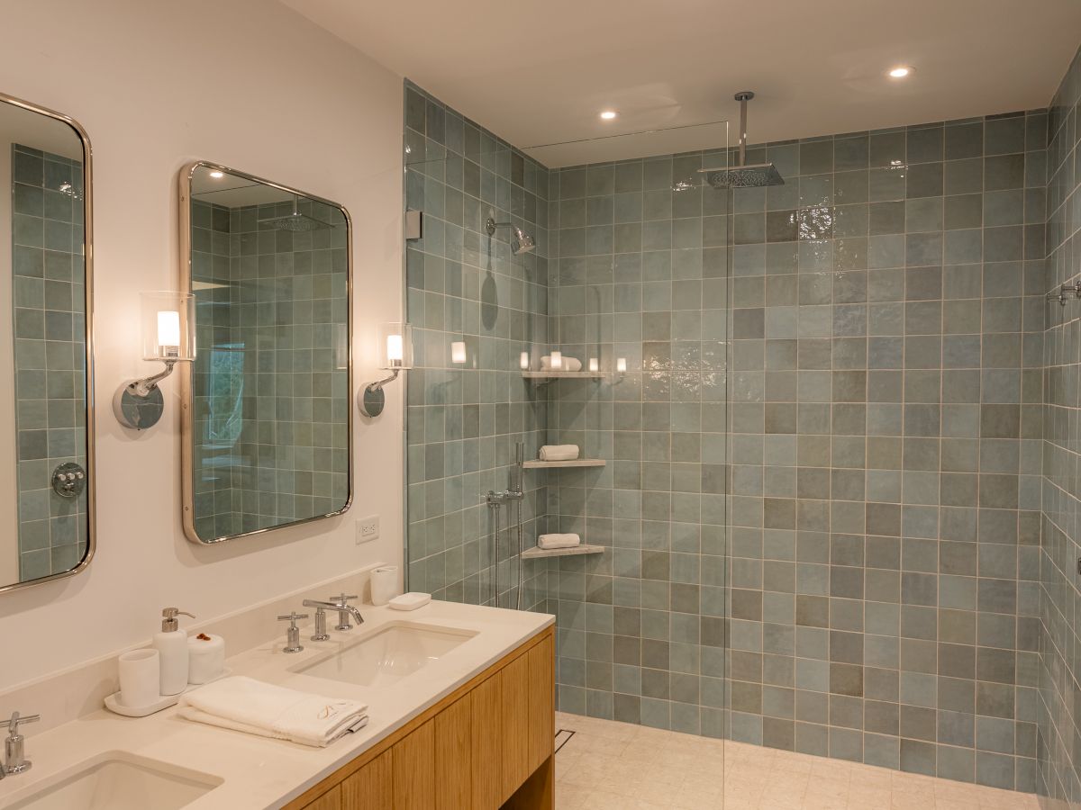 A sleek bathroom with a double sink vanity, two wall-mounted mirrors, sconces, and a spacious tiled walk-in shower in soft blue-green tiles.