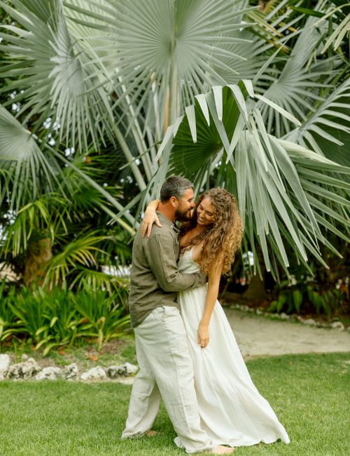 A couple dancing closely in a garden, eucalyptus-like fan palms overhead, green foliage around, a romantic outdoor scene.