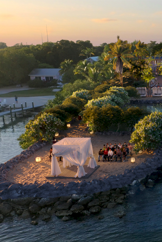 A tropical waterfront scene at dusk with a small stone jetty, white tent, group dining under string lights, palms, and a resort backdrop.