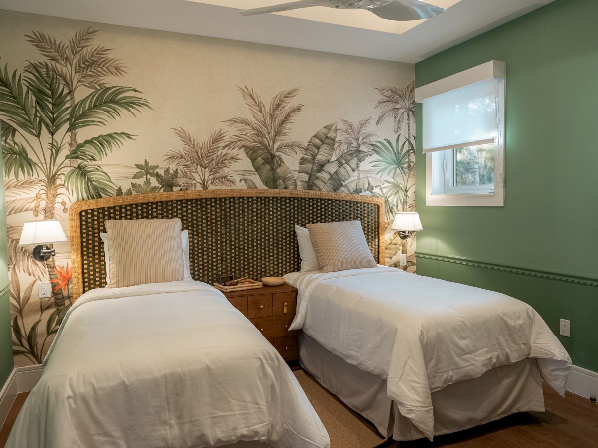 Two twin beds with white linens and pillows, a shared wooden headboard, nightstands with lamps, tropical palm wallpaper, green walls, and a window with a shade.