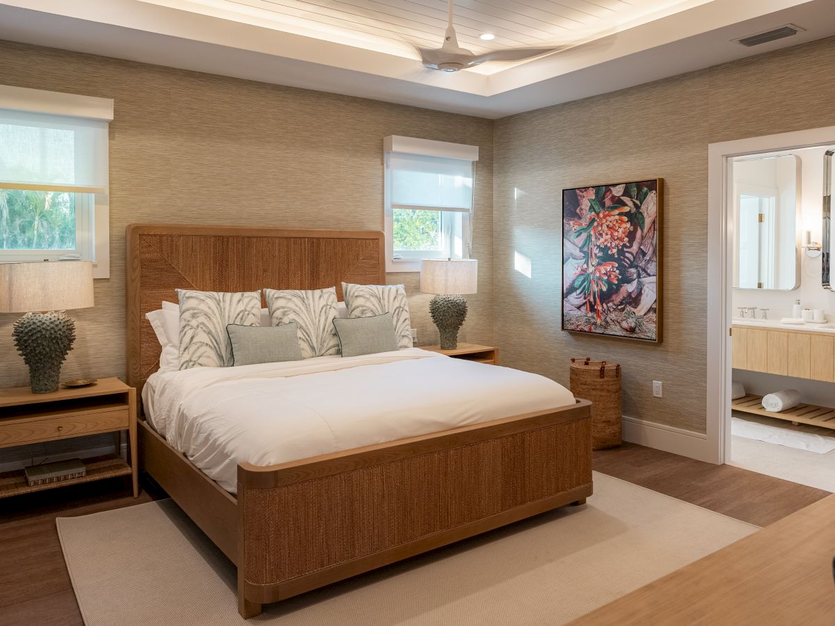 A modern hotel bedroom with a wooden bed, two nightstands, two lamps, soft neutral walls, a ceiling fan, and a doorway to a bright en-suite bathroom.