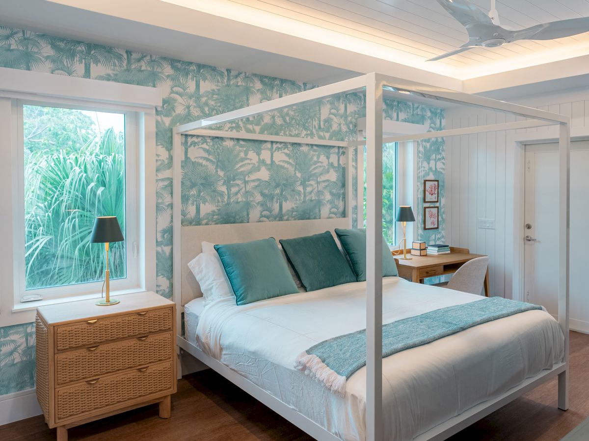 A bright, tropical-themed hotel bedroom with a white four-poster bed, teal accents, palm wallpaper, wooden nightstand, and a window view.