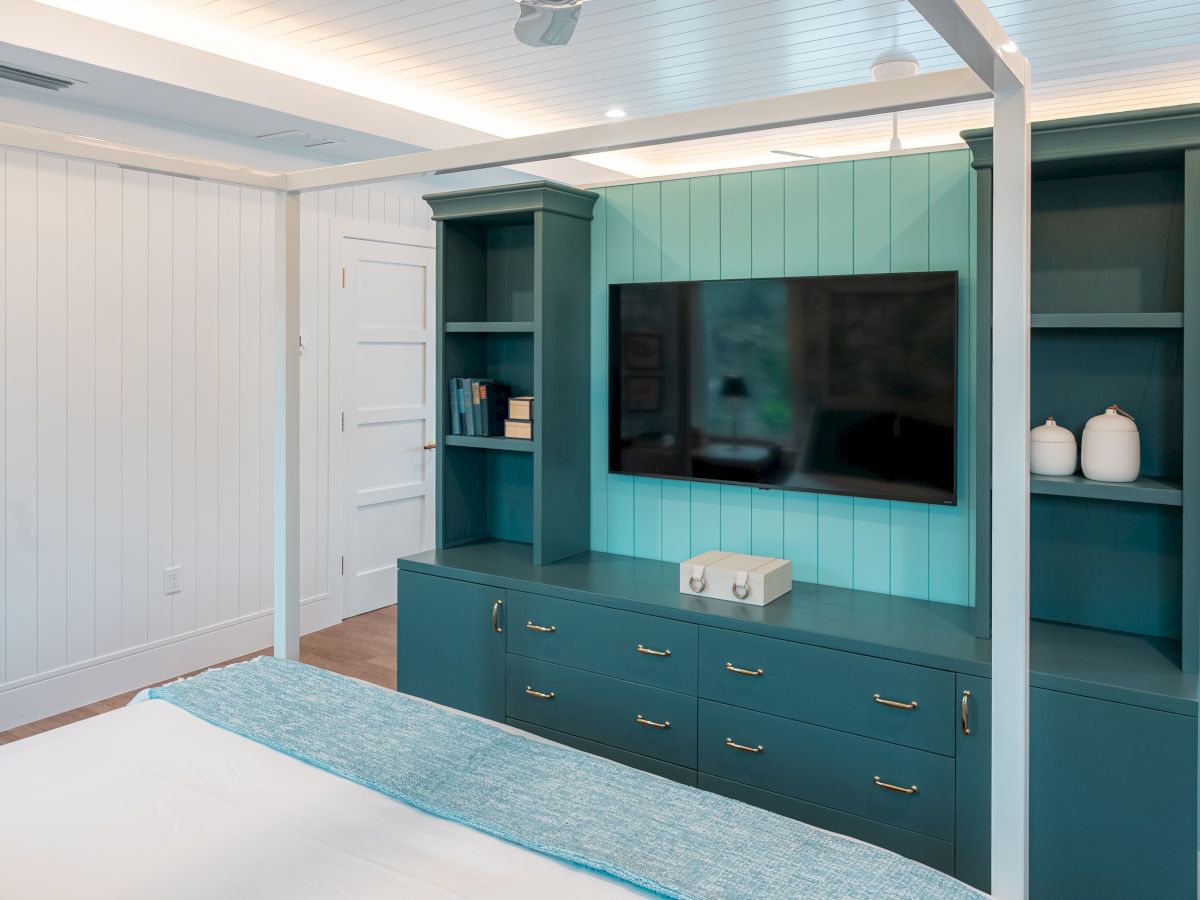 A teal built-in media wall with a mounted TV, white cabinet drawers, open shelves, and decorative jars, forming a cozy entertainment setup in a bright room.