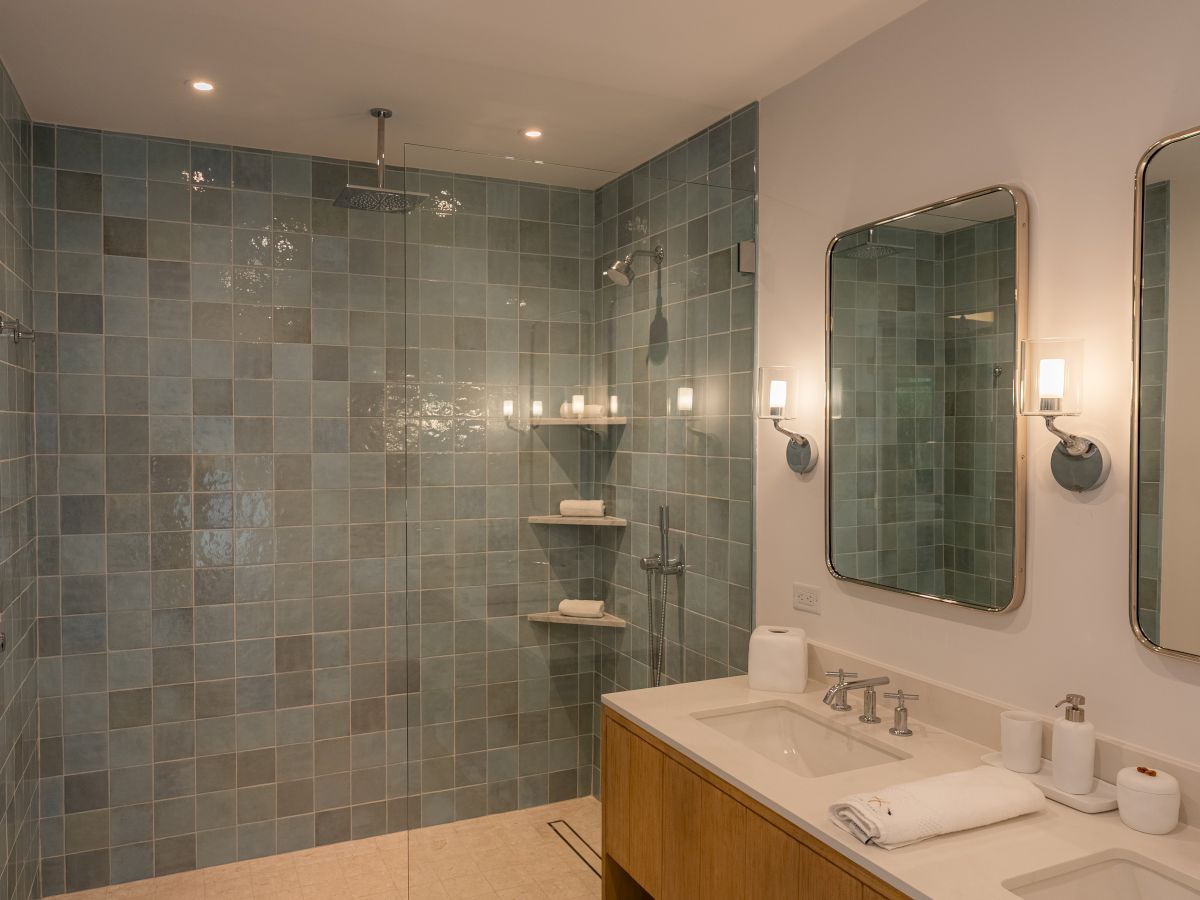 A modern bathroom with a blue tile walk-in shower, double vanity, and two wall-mounted mirrors with sconces, plus towels and toiletries.