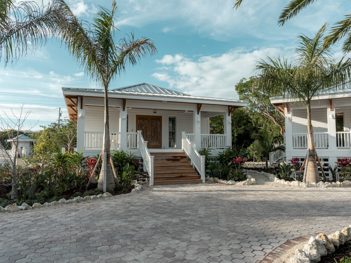 A row of small, white seaside cottages with wooden steps, palm trees, and a paved courtyard under a bright, sunny sky.