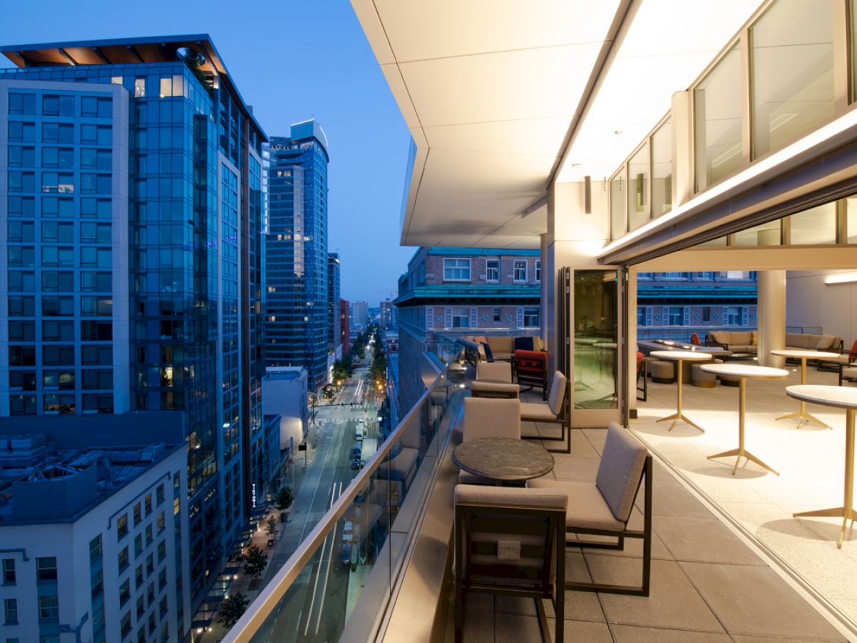 A modern rooftop patio with tables and chairs overlooks a cityscape of tall buildings at dusk.