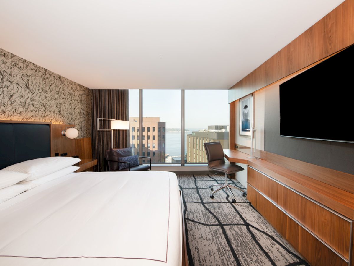 A modern hotel room with a large bed, wall-mounted TV, desk, chair, and a window offering a cityscape view.