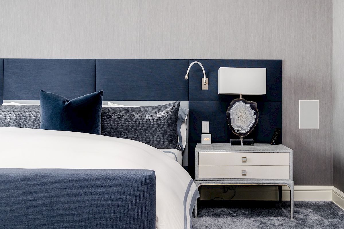 A modern bedroom with a blue headboard, white and blue bedding, and a nightstand featuring a lamp and decorative object.