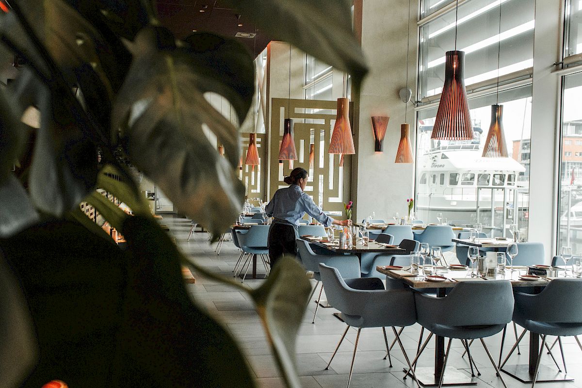 A person setting tables in a modern restaurant with plants and stylish lighting features, creating a cozy ambiance.
